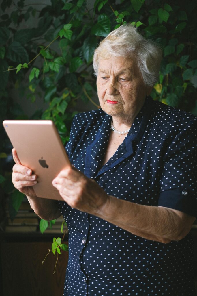 Aged serious female with wrinkles in dress surfing contemporary tablet near plant with green leaves