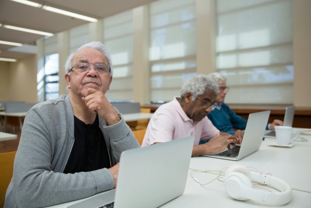 Three senior men engaged with laptops in a library environment, promoting lifelong learning.